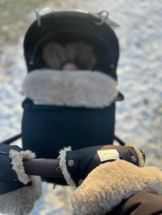 A person's hand holding a MellowConceptStore Waterproof Natural Sheepskin Stroller Hand Muffs - Black&White in the snow.