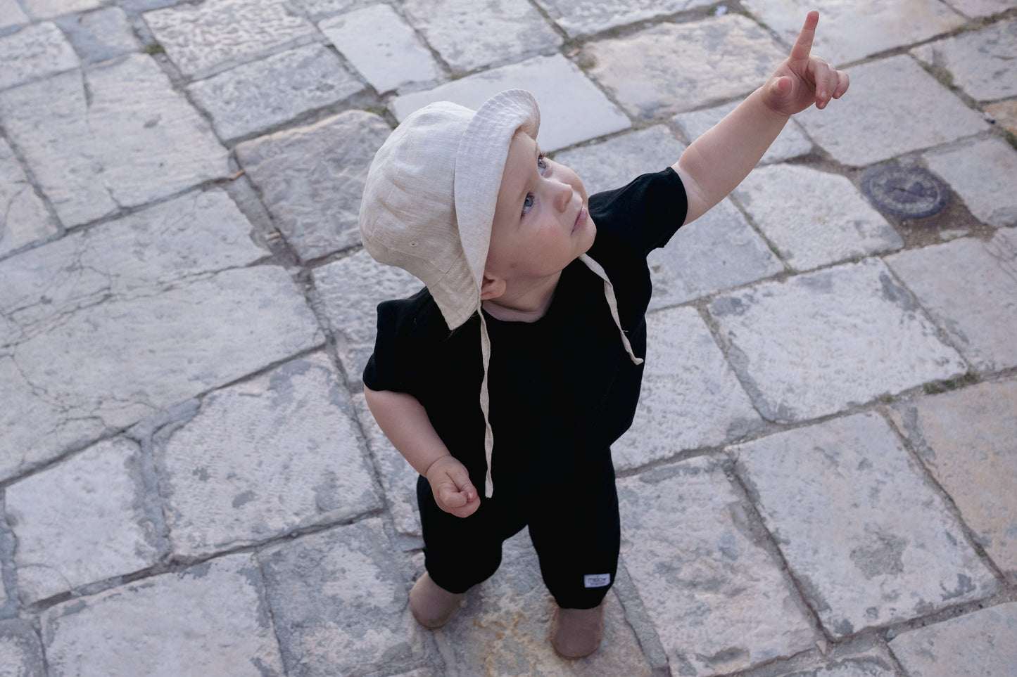A baby boy pointing at something on a cobblestone street, wearing the Ramie Baby/Kid Clothing Set in Charcoal by Mellow Concept Store.