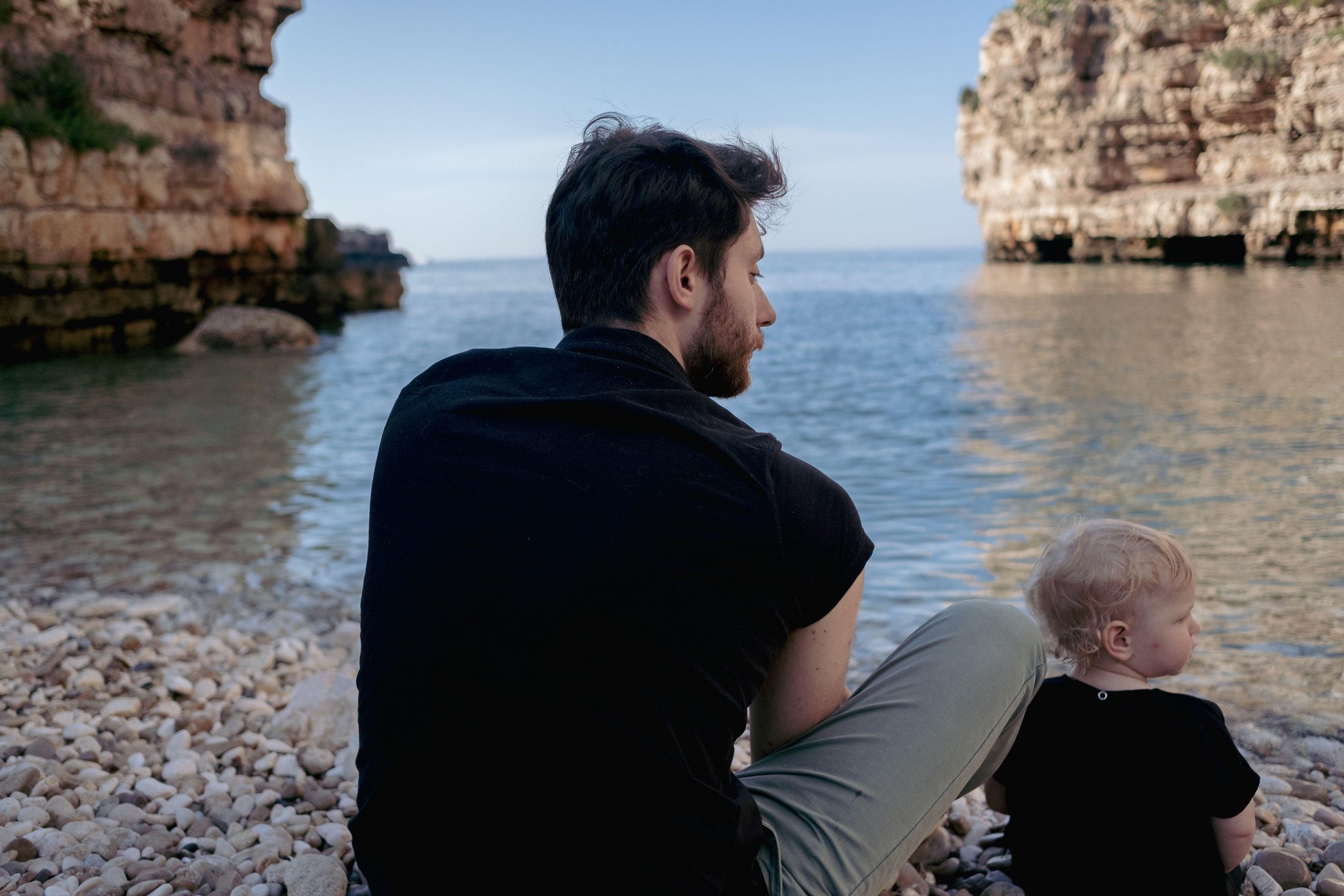 A man and a child sitting on the beach, gazing at the ocean, both wearing Mellow Concept Store's Ramie Baby/Kid Clothing Set in Charcoal.