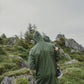A person wearing a Khaki MellowConceptStore waterproof baby/kid clothing set with a hood stands in a mountainous landscape, facing towards the forest-covered slopes under a cloudy sky.