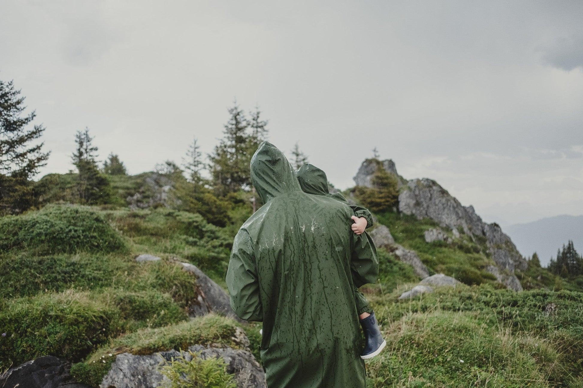 A person wearing a Khaki MellowConceptStore waterproof baby/kid clothing set with a hood stands in a mountainous landscape, facing towards the forest-covered slopes under a cloudy sky.