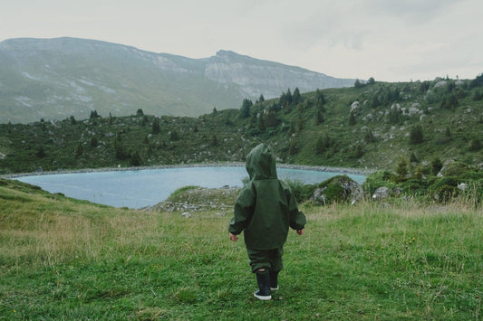 A child in a Waterproof Baby/Kid Clothing Set - Khaki from MellowConceptStore stands on a grassy field, facing a mountainous landscape with a clear blue lake.