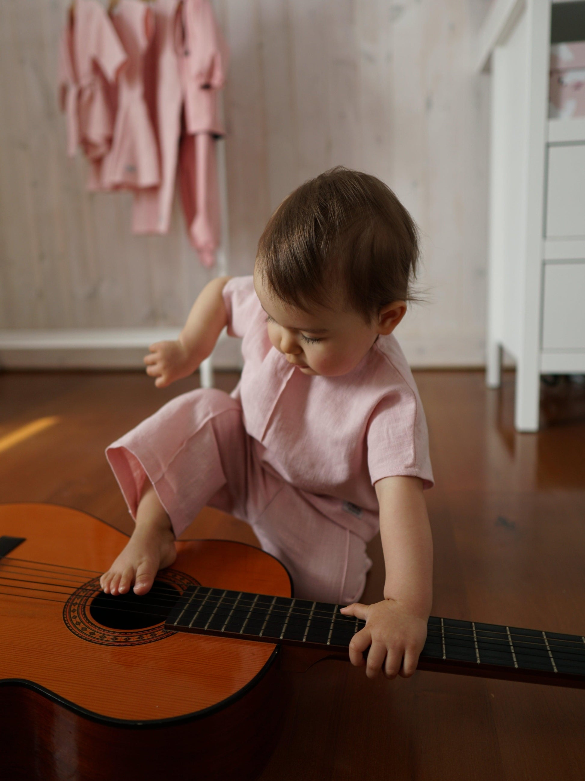 A baby strums on a Ramie Baby/Kid Clothing Set in Rosa, perfect for delicate skin from Mellow Concept Store.