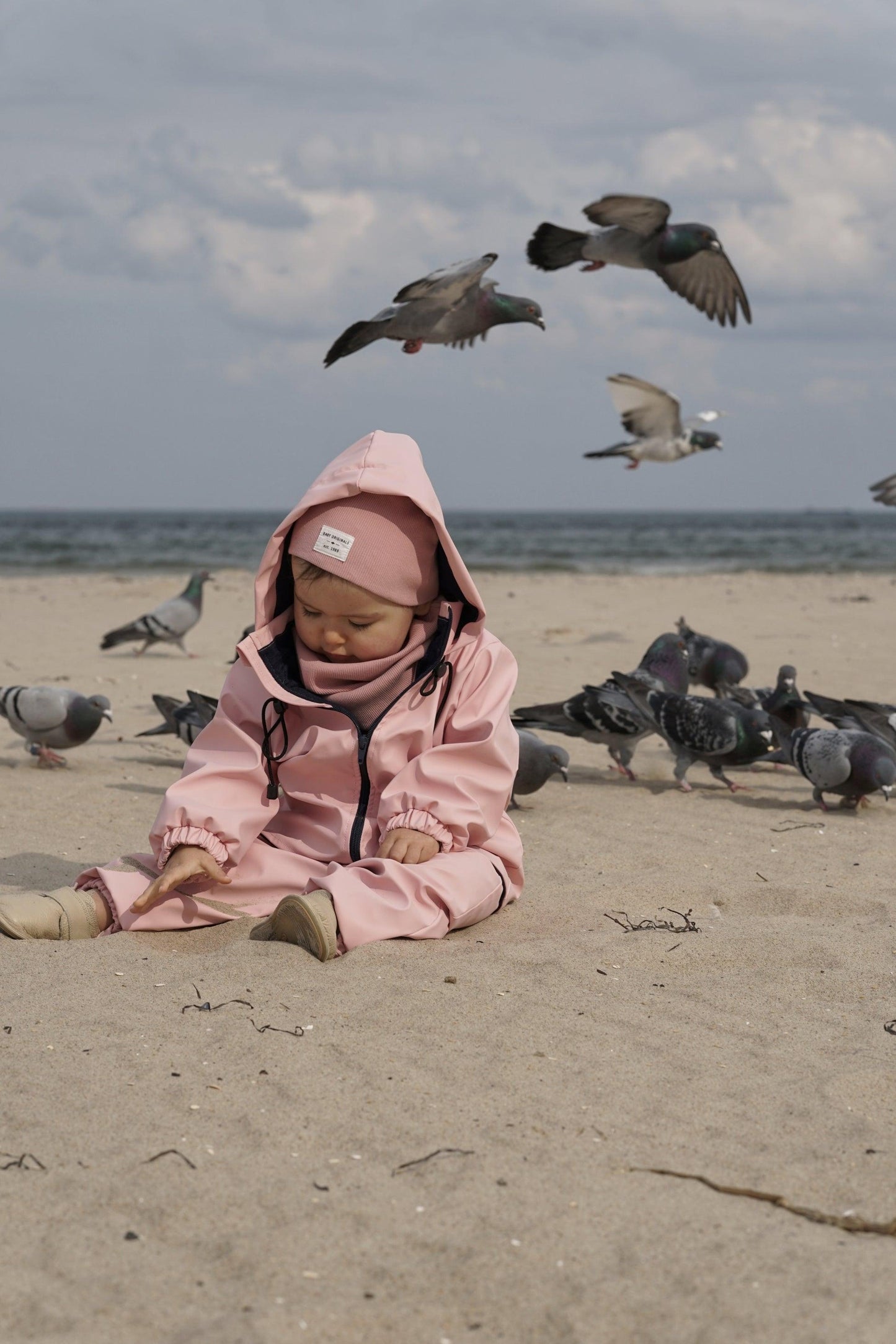 A child in a pink jacket with adjustable suspenders sits on the sand surrounded by pigeons, with some birds in flight near them wearing the Waterproof Baby/Kid Clothing Set - Rosa from MellowConceptStore.