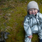 A smiling child wearing a MellowConceptStore Waterproof Baby/Kid Clothing Set in Silver and a white merino wool beanie sits on a green mossy ground.