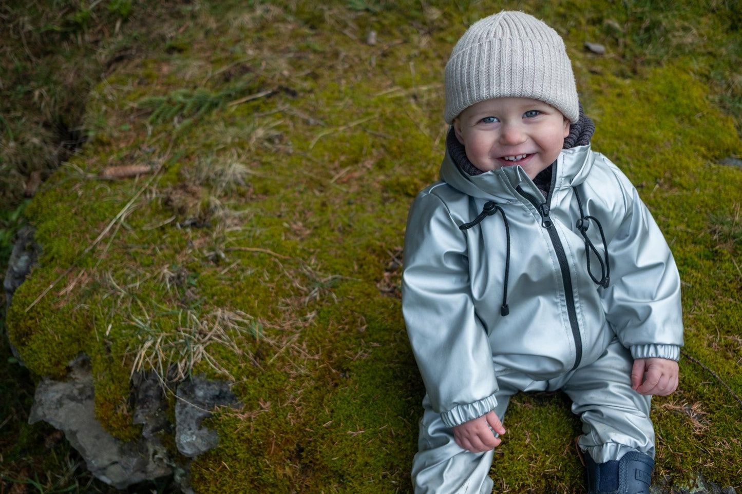 A smiling child wearing a MellowConceptStore Waterproof Baby/Kid Clothing Set in Silver and a white merino wool beanie sits on a green mossy ground.