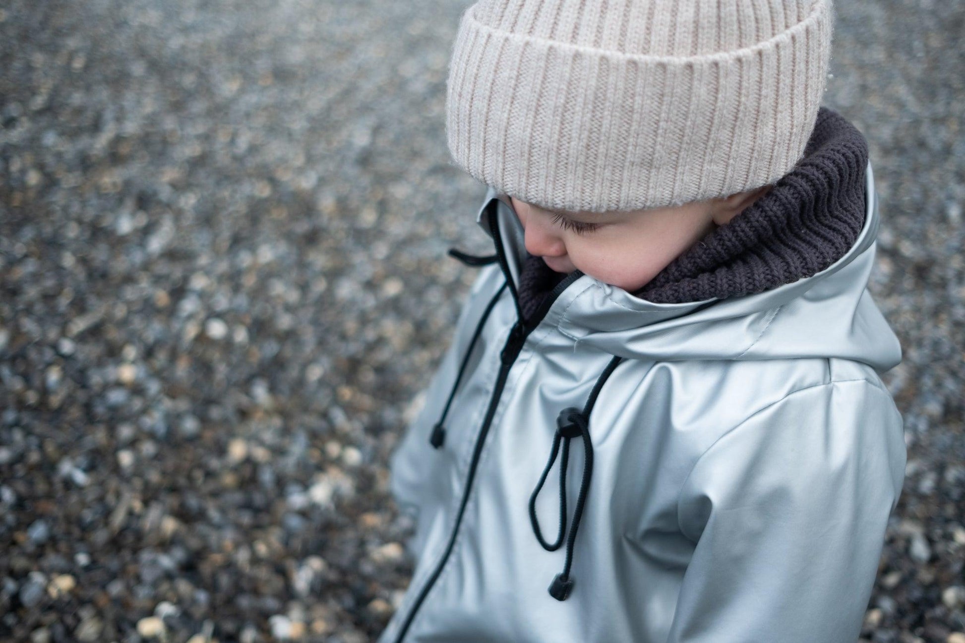 A child dressed in warm, Waterproof Baby/Kid Clothing Set - Silver from MellowConceptStore, including a hat and a hooded coat made of merino wool, is looking downwards against a background of small stones or gravel.