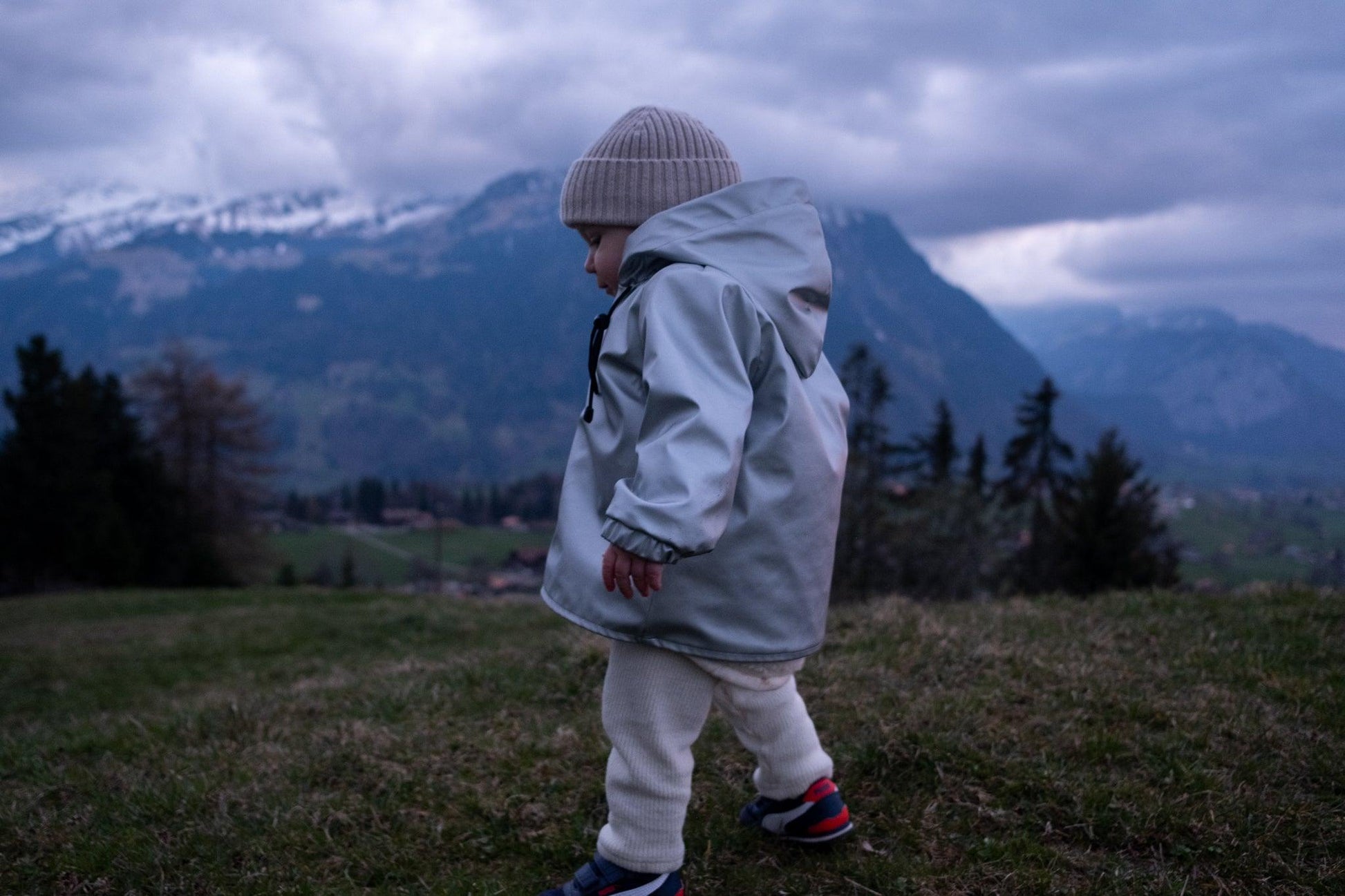 A toddler wearing a white merino wool beanie and a grey MellowConceptStore Waterproof Baby/Kid Clothing Set - Silver jacket is walking on grass with mountains visible in the background during dusk.