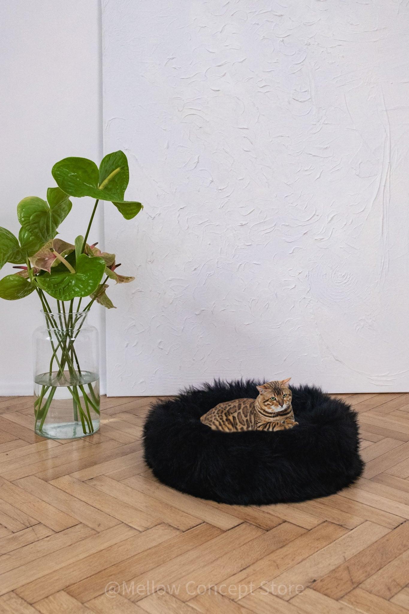 A black cat relaxing on a fluffy Round Natural Sheepskin Pet Bed in front of a potted plant from Mellow Pet Store.