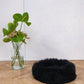 A black Round Natural Sheepskin Pet Bed made by Mellow Pet Store on a wooden floor.