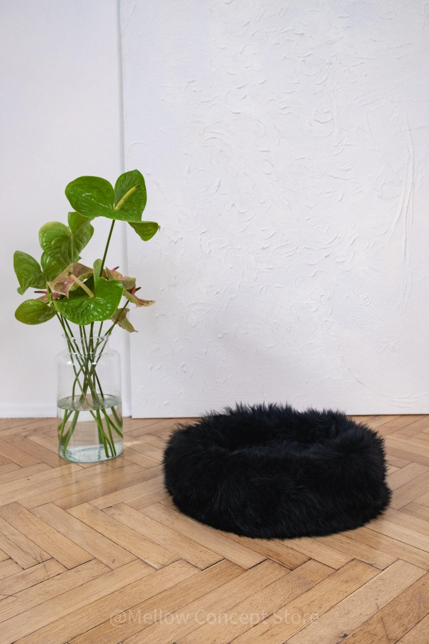 A black Round Natural Sheepskin Pet Bed made by Mellow Pet Store on a wooden floor.
