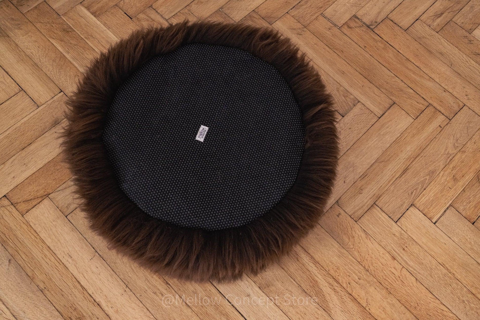 A black fur ball lounging on a wooden floor with Round Natural Sheepskin Pet Bed in Brown from Mellow Pet Store nearby.