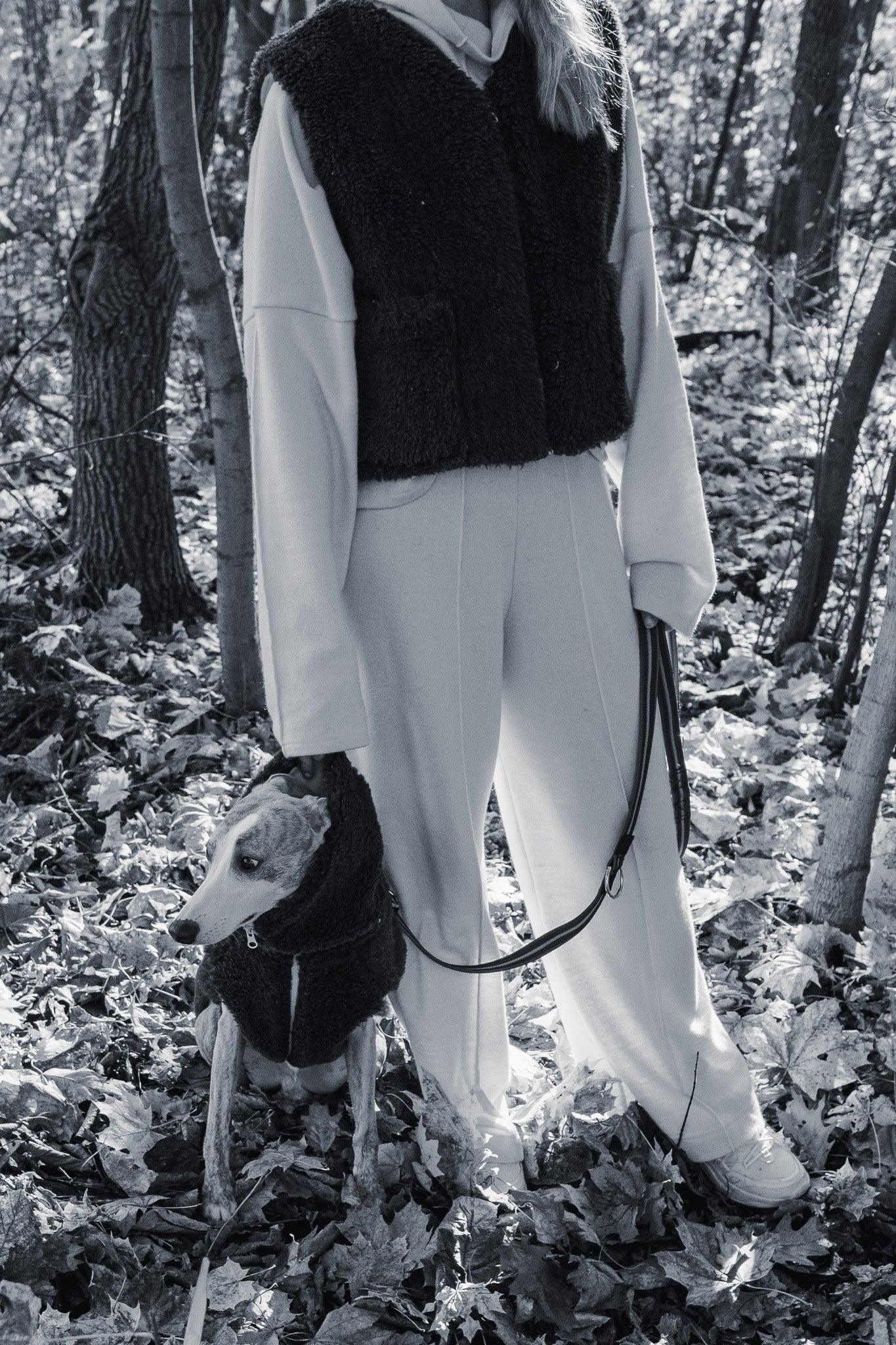 A woman standing in the woods with a dog on a leash, enjoying nature, wearing a Woolen Dog Vest in Charcoal by Mellow Pet Store.
