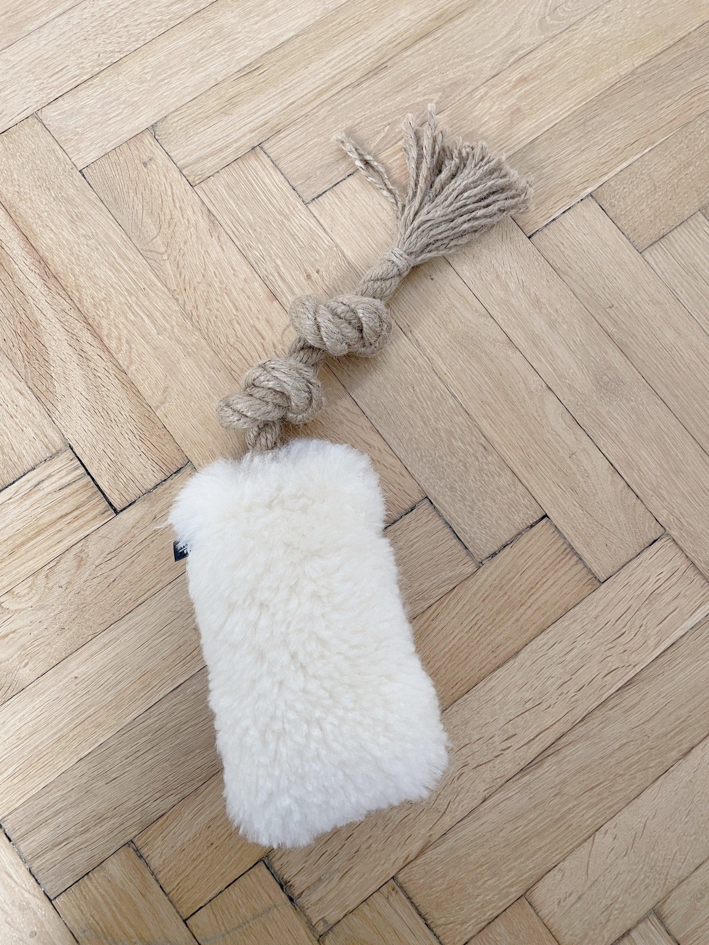A white Mellow Pet Store sheepskin dog tug toy with a tassel rests on the wooden floor. Crafted from natural sheepskin, this pet accessory exudes luxury and comfort.